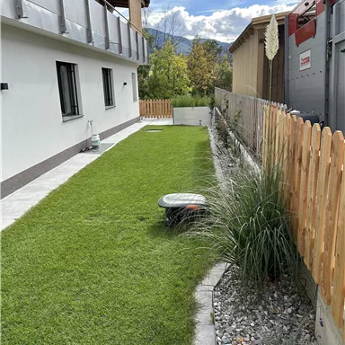 Ein gepflegter Garten mit grünem Rasen und modernen Pflanzen. Im Hintergrund sind ein Haus und ein Sichtschutz aus Holz zu sehen.