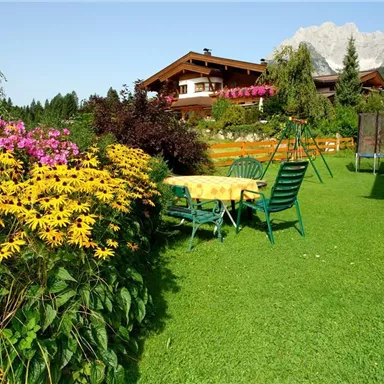 Ein schöner Garten mit bunten Blumen und einem Tisch mit Stühlen. Im Hintergrund ist ein gemütliches Haus und die Berge sind sichtbar.