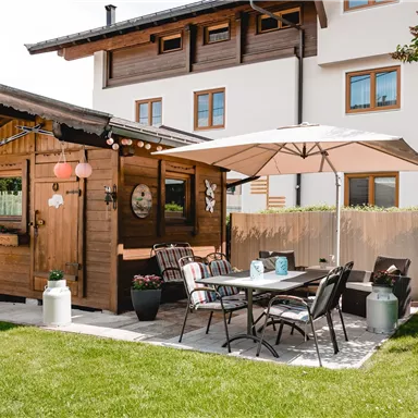 Ein rustikales Gartenhaus aus Holz mit einem gemütlichen Sitzbereich. Der Garten ist gepflegt und hat Rasen, Tische und Stühle unter einem Sonnenschirm.