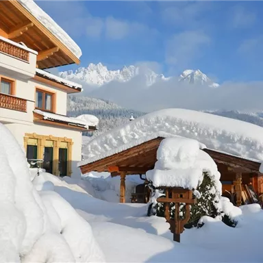 A snow-covered house in an idyllic winter landscape. The mountains in the background are clear and the sun is shining.