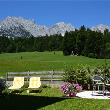 Eine malerische Landschaft mit grünen Wiesen und majestätischen Bergen im Hintergrund. Auf der Terrasse stehen Sonnenliegen und Gartenmöbel.