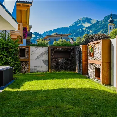 Ein schöner Garten mit grünen Rasenflächen und modernen Steinmauern. Im Hintergrund sind Berge und ein klarer blauer Himmel zu sehen.