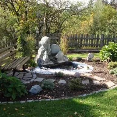 A small, idyllic garden landscape with a pond and a stone installation. A wooden bench invites you to relax and enjoy nature.