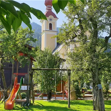 Ein schöner Garten mit Spielplatz und Bäumen. Im Hintergrund ist eine Kirche mit einem roten Turm zu sehen.