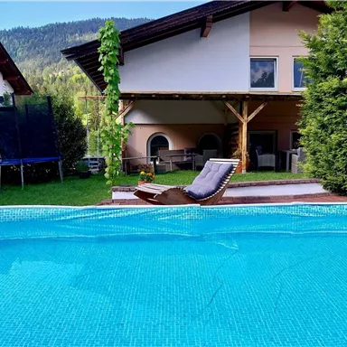 A beautiful pool with clear water and a sun lounger beside it. In the background, there is a house with a covered area and plenty of greenery.