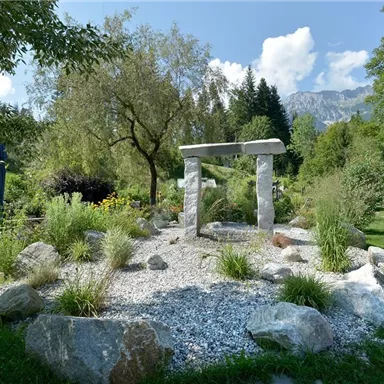 A tranquil garden with a stone installation and many plants. In the background, trees and mountains can be seen.