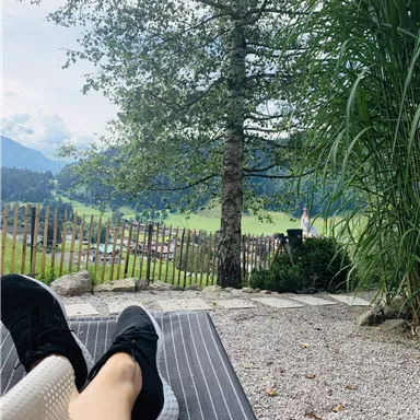 A relaxed lounging position in nature with a view of green meadows and mountains. A tree and tall plants frame the scene.