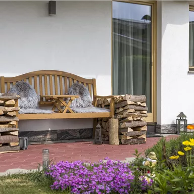 A cozy bench with pillows, surrounded by a well-maintained garden. Next to the bench lies a stack of wood and colorful flowers are blooming.