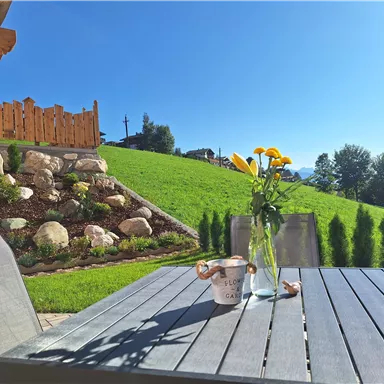 Ein schöner Außenbereich mit einem Tisch und einer Vase voller gelber Blumen. Im Hintergrund sind grüne Wiesen und gepflegte Landschaftselemente zu sehen.