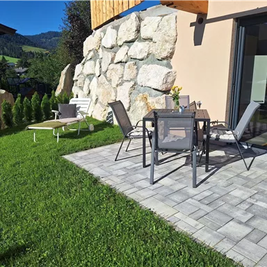 Ein schöner Außenbereich mit einem gepflasterten Boden und Gartenmöbeln. Im Hintergrund sind grüne Pflanzen und eine Felsenwand zu sehen.