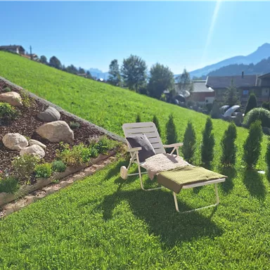Ein ruhiger Garten mit grünem Gras und Sonnenliegen. Im Hintergrund sind Hügel und Bäume zu sehen.