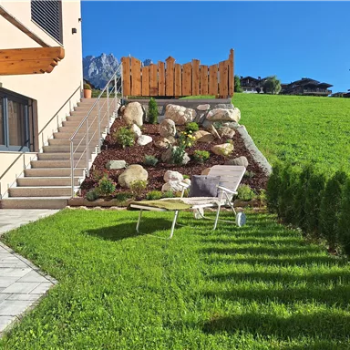 Ein gepflegter Garten mit grünem Rasen und einem bequemen Liegestuhl. Im Hintergrund sind Felsen und eine Holzumzäunung zu sehen.