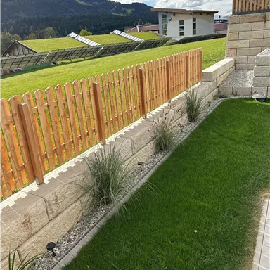 Ein gepflegter Garten mit grünem Rasen und einem Holzlattenzaun. Im Hintergrund sind Berge und Gebäude sichtbar.