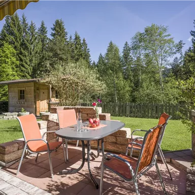 Eine gemütliche Terrasse mit einem Tisch und Stühlen, umgeben von grünen Bäumen. Der Himmel ist blau und klar, ideal für entspannte Stunden im Freien.