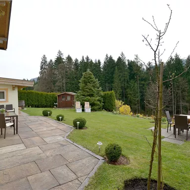 Ein schön gestalteter Garten mit gepflastertem Weg und Sitzgelegenheiten. Im Hintergrund sind hohe Bäume und ein ruhiger Ausblick zu sehen.