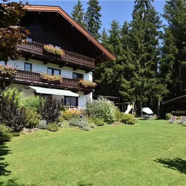 Ein hübsches Haus umgeben von einem gepflegten Garten. Im Hintergrund sieht man hohe Bäume und einen sonnigen Himmel.