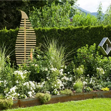Ein schöner Garten mit einer blühenden Pflanzenvielfalt und einer dekorativen Holzskulptur. Im Hintergrund sind hohe Pflanzen und eine grüne Hecke zu sehen.
