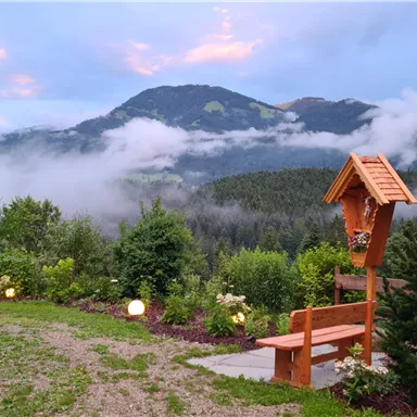 Eine ruhige Landschaft mit Bergen im Hintergrund und sanften Wolken. Im Vordergrund steht eine Bank, umgeben von Bäumen und Gartenbeleuchtung.