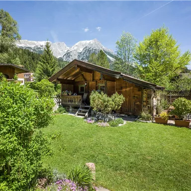 Ein rustikales Holzhaus in einem grünen Garten, umgeben von Bäumen und Blumen. Im Hintergrund sind schneebedeckte Berge zu sehen.