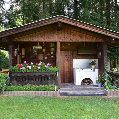 Ein gemütliches Holzhaus mit bunten Blumen im Vordergrund. Es steht in einer grünen Umgebung, umgeben von Bäumen.