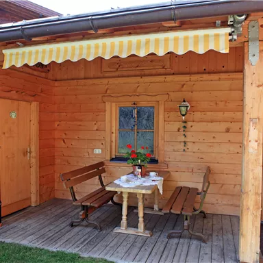 An inviting outdoor area with a wooden porch and a small table. Two benches and an awning create a cozy atmosphere.
