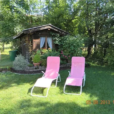Ein schöner Garten mit zwei pinken Liegestühlen. Im Hintergrund steht eine kleine Holzhütte, umgeben von grüner Vegetation.