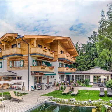 Ein schönes Hotel im alpenländischen Stil mit Holzverkleidung. Der Garten ist grün und einladend, mit Liegestühlen und einem Schwimmbecken.