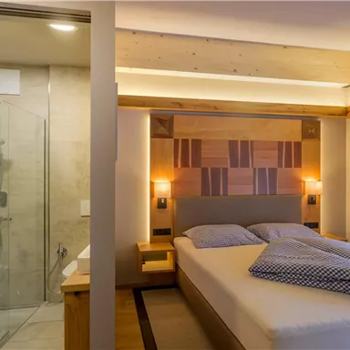 Ein modernes Zimmer mit einem Doppelbett und stilvollen Lichtquellen. Im Hintergrund ist eine Duschkabine aus Glas zu sehen.