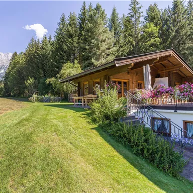 Ein gemütliches Holzhaus umgeben von Bäumen und Grünflächen. Im Hintergrund sind Berge sichtbar, was eine malerische Landschaft schafft.