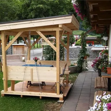 Eine gemütliche Holzlauben im Garten mit einem Tisch und Stühlen. Blumen und Pflanzen schmücken den Bereich um die Laube.
