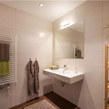 Ein modernes Badezimmer mit hellen Fliesen und einem großen Spiegel. Handtücher und ein Regal mit Pflegeartikeln vervollständigen die Einrichtung.