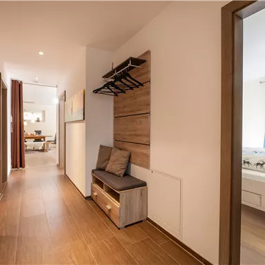 Ein heller Flur mit Holzfußboden und einer Sitzgelegenheit. Auf der linken Seite befinden sich Kleiderhaken und der Zugang zu einem Wohnzimmer.