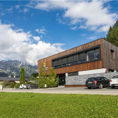Ein modernes Gebäude mit Holzelementen und großen Fenstern steht in einer malerischen Landschaft. Im Hintergrund sind Berge und ein klarer Himmel zu sehen.