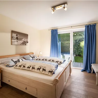 Ein helles Schlafzimmer mit einem großen Doppelbett und Holzmöbeln. Vor den Fenstern hängen blaue Vorhänge, und es gibt einen Blick nach draußen in den Garten.