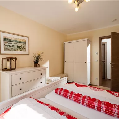 Ein helles Schlafzimmer mit zwei Einzelbetten und einer rot-weißen Decke. An der Wand hängt ein Bild, und es gibt einen weißen Schrank sowie eine Holzkommode.