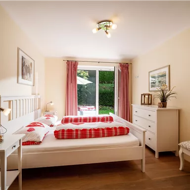 Ein gemütliches Schlafzimmer mit einem Doppelbett, roten Dekokissen und einem Schreibtisch. Große Fenster sorgen für viel Tageslicht und einen Blick ins Grüne.