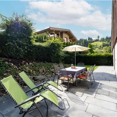 Ein einladender Patio mit einem Tisch und Stühlen, umgeben von grünen Pflanzen. Im Hintergrund ist ein Holzhaus und eine hügelige Landschaft zu sehen.