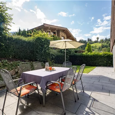 Ein gemütlicher Außensitzbereich mit einem Tisch und Stühlen unter einem Sonnenschirm. Im Hintergrund sind grüne Hügel und ein attraktives Haus zu sehen.