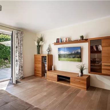 Ein modernes Wohnzimmer mit holzfarbenen Möbeln und großem Fenster. Im Hintergrund sieht man eine grüne Gartenlandschaft.