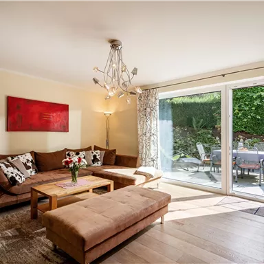 Ein heller Wohnraum mit einem gemütlichen Sofa und einem Holztisch. Große Fenster bieten einen Blick auf einen Gartenbereich.