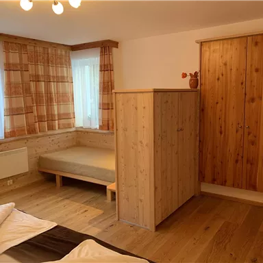 Ein einfaches Schlafzimmer mit Holzmöbeln und einem Bett. Die Wände sind hell und es gibt große Fenster mit Vorhängen.