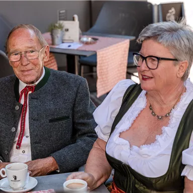 An older couple sits in traditional attire in a café. They smile and enjoy their drinks in a cozy atmosphere.
