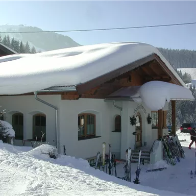 Ein charmantes Haus im Schnee mit einem schneebedeckten Dach. Im Vordergrund stehen Skier, und die Umgebung ist winterlich und idyllisch.