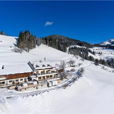 Ein gemütliches Haus inmitten einer verschneiten Landschaft. Im Hintergrund erstrecken sich schneebedeckte Hügel und ein blauer Himmel.