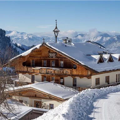 Ein traditionelles Holzhaus im Winter mit Schnee bedeckt. Im Hintergrund sind verschneite Berge und ein blauer Himmel zu sehen.