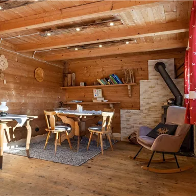 Ein gemütlicher Raum mit Holzwänden und einem Tisch, umgeben von Stühlen. Ein Kamin und Regale mit Büchern verleihen dem Raum eine einladende Atmosphäre.