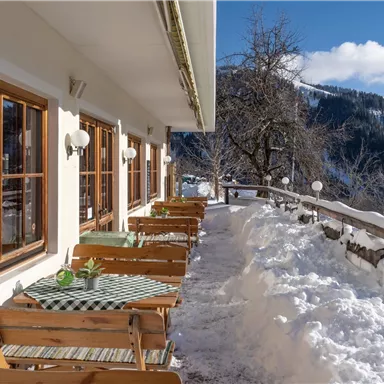 Eine gemütliche Terrasse eines Restaurants mit Holzstühlen und Tischen, umgeben von Schnee. Im Hintergrund sind schneebedeckte Berge und ein blauer Himmel zu sehen.