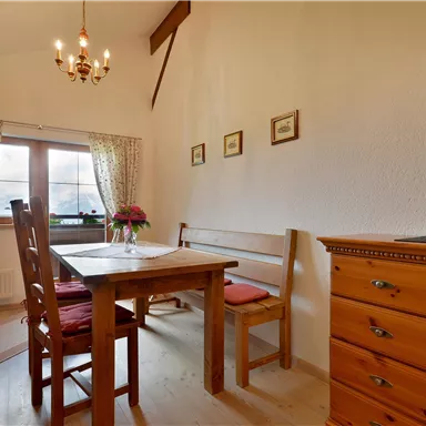 A cozy dining area with a wooden table, chairs, and blooming plants on the windowsill. The walls are in a warm shade, and there is a beautiful ceiling light.