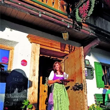 Eine fröhliche Frau in traditioneller Tracht hält ein Tablett vor einem gemütlichen Gasthaus. Die Fassade des Gebäudes ist mit Blumen und einer einladenden Atmosphäre dekoriert.