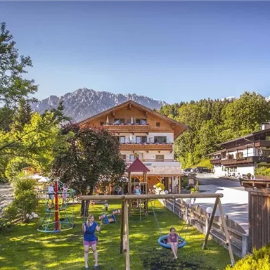 A picturesque mountain village with traditional wooden houses and green meadows. In the foreground, there is a playground and the impressive mountains in the background.